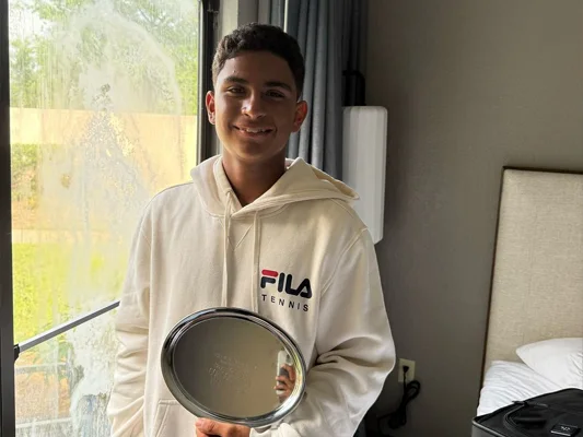 An adolescent boy smiling with a silver tennis tournament trophy.